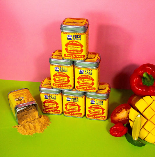 Stack of mango habanero seasonings with a red pepper and pineapple on a green surface against a pink background